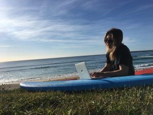 Henkilö istuu meren rannalla surffilaudan päällä ja kirjoittaa tietokoneella.