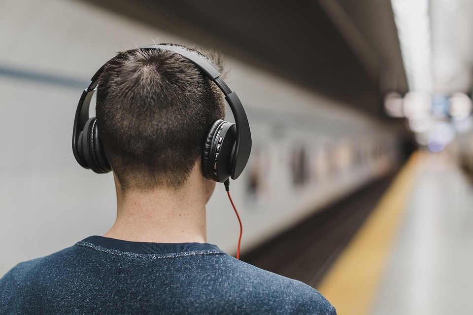 Lähikuva asemalaiturilla olevasta miehestä, jolla on kuulokkeet korvilla. Mies on selin katsojaan. Taustalla näkyy juna. 