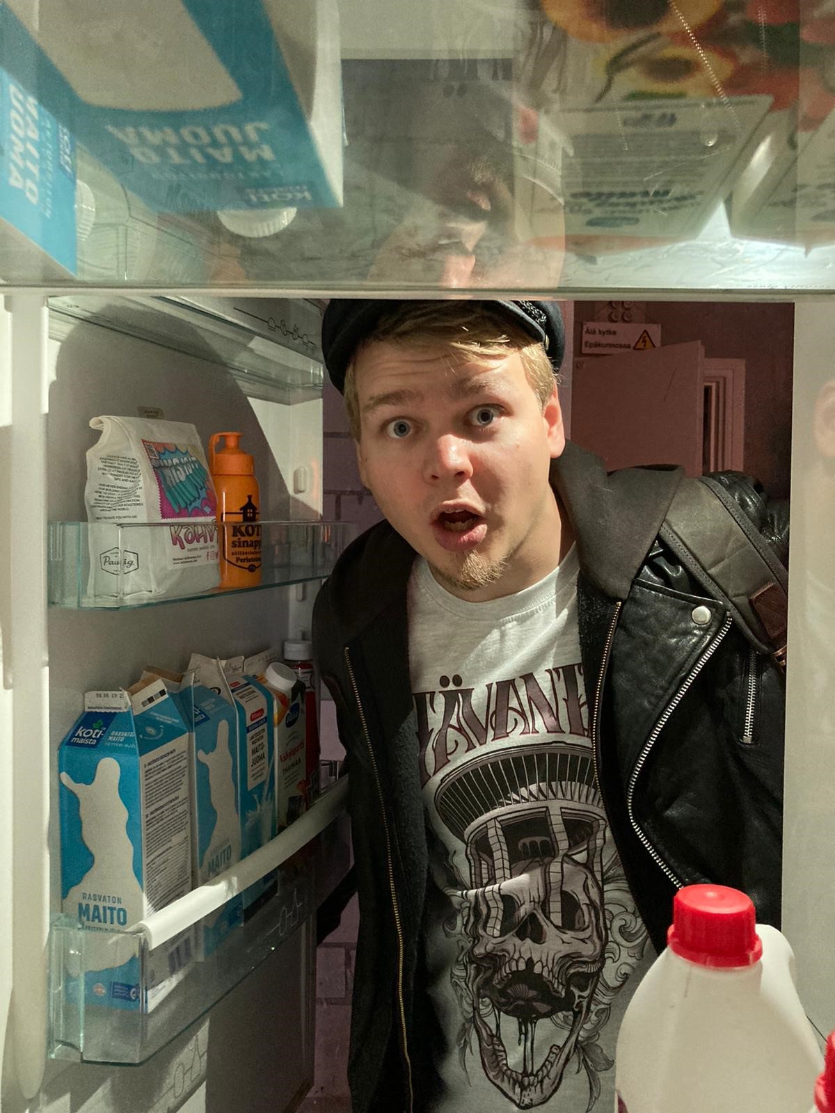 Ilmoisen hämmästynyt mies lakki päässä kurkistaa jääkaapin ovelta, maitopurkkeja on jääkaapin ovessa.