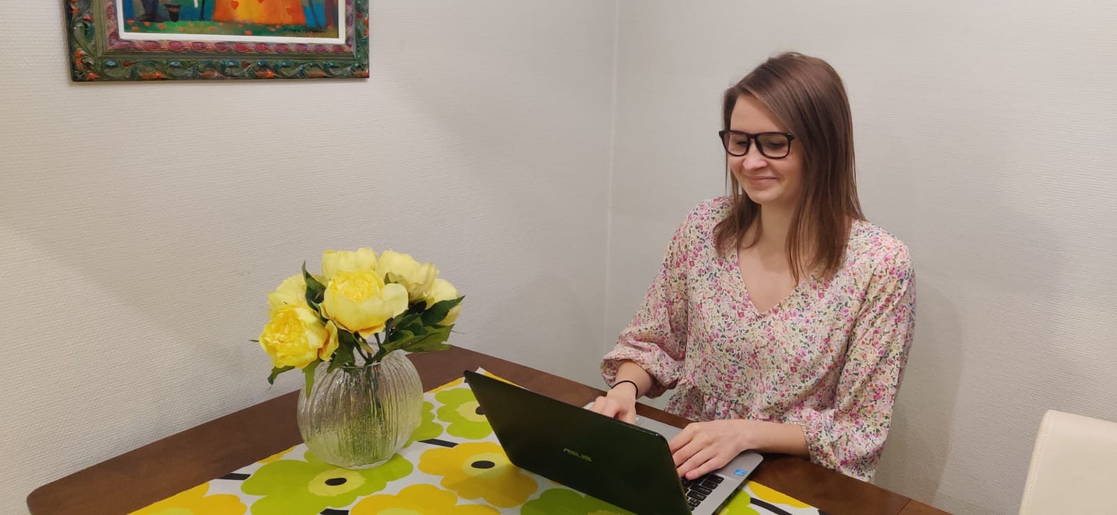 Nainen istuu tietokoneen ääressä ja pöydällä on keltaisia kukkasia.