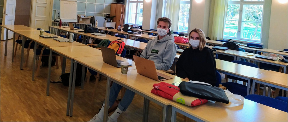Luokkatilassa istuvat pulpettirivistön keskellä mies ja nainen maskit päällä, taustalla ikkunat ja pöydillä opiskelijoiden tavaroita.