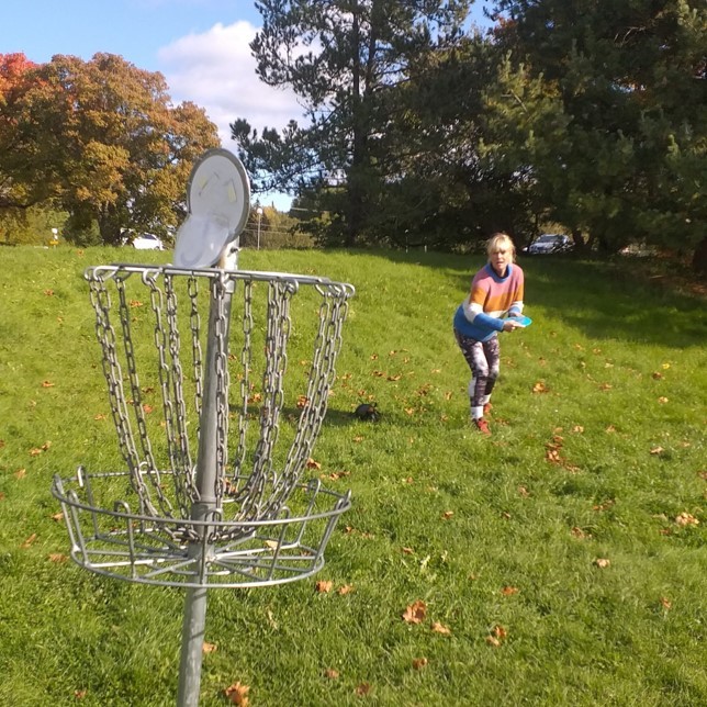 Nainen heittää frisbeegolf-kiekkoa kohti koria, joka on noin 5 metrin päässä, aurinkoisella nurmikolla.