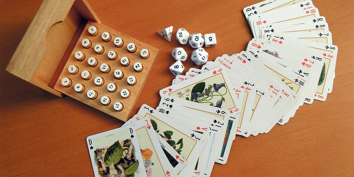 Blogi Nuorisotyöntekijästä pelikasvattajaksi: Hajallaan oleva korttipakka, jonka vieressä on erimuotoisia noppia ja puinen yksinpelattava lautapeli.
