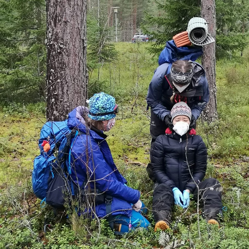 Three persons in a finnish forest. One has had an accident and is sitting on the ground, one is holding that persons head stable hands on the sides of the head and one is talking to a patient. 