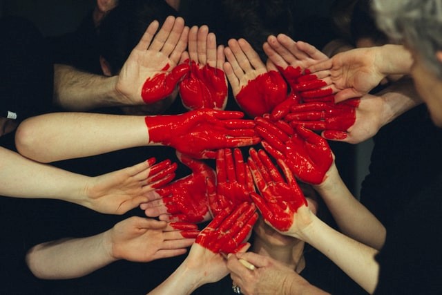Seitsemän ihmistä ovat ojentaneet kämmenensä ilmaan vierekkäin. Kämmenien muodostamaan  alustaan on maalattu punainen iso sydän.