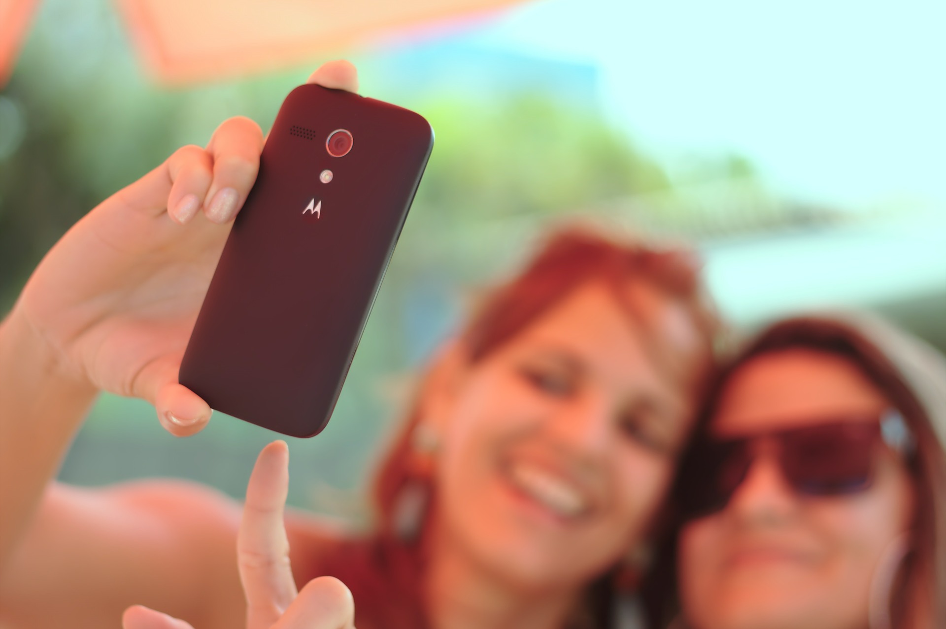 Kuvassa taustalla kaksi hämyisää nuorta naista, jotka ottavat selfietä, kuva tarkentuu etuosassa näkyvään kännykkään ja siitä kiinni pitävään käteen. 