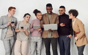 Six people from different cultures are standing next to one another. They all have gadgets in their hands and all are looking over at one of the people's laptop.