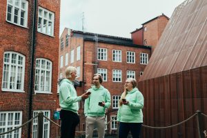 Commu-sovelluksen kolmihenkinen perustajatiimi seisoo hymyillen ja keskustellen vihreissä Commu-huppareissa ja älypuhelimet kädessä rakennuksien edessä.