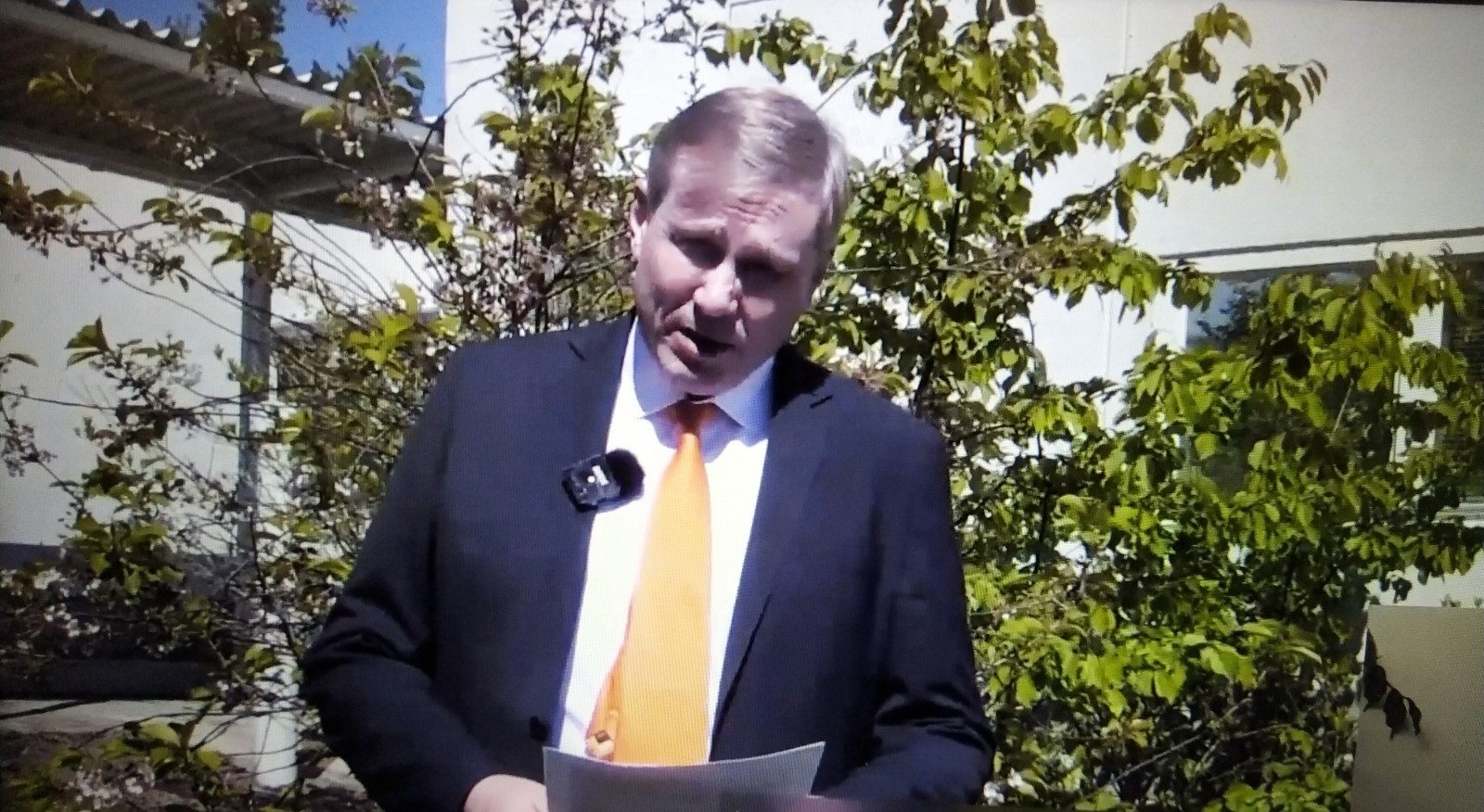 Jukka Määttä seisoo talojen ja lehtipuiden edessä pihalla pitämässä puhetta paperit käsissään.