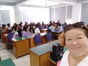Naisen ottama selfie, jonka taustalla näkyy luokkahuone täynnä työskenteleviä opiskelijoita.