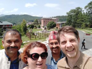 A selfie of four people standing outside in the sun - three men and one woman. There are buildings, a fountain and mountains in the background.
