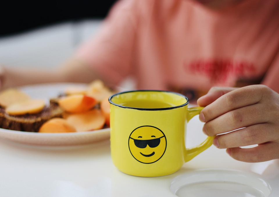 Kuvassa keltainen muki, jossa on hymyilevä emoji, jolla on aurinkolasit. Mukin takana on lautanen, jossa on ruokaa.