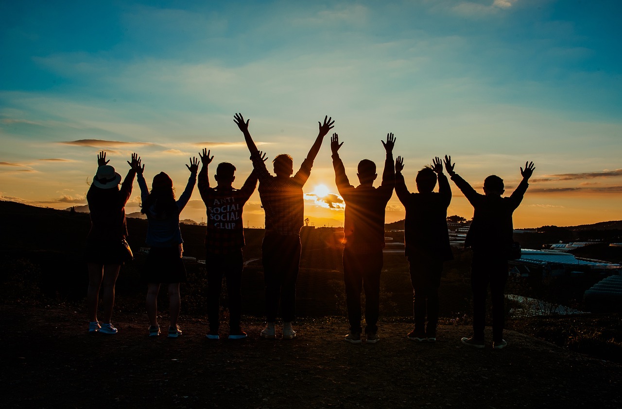 : seven young people standing side by side watching the sunset together, arms raised towards the sky
