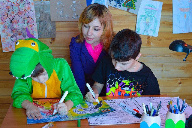 Vaihtoehtoinen teksti: Kaksi lasta värittävät värityskuvaa. Heidän välissää istuu nainen, joka katsoo poikien työskentelyä.