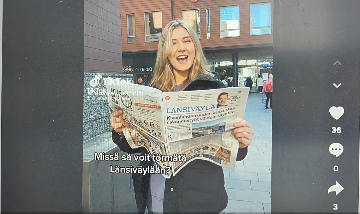 Pitkähiuksinen nainen seisoo ulkona ja hänellään on kädessään auki oleva Länsiväylä-lehti. Taustalla tiilirakennus. Kuvakaappaus tiktok-videosta.