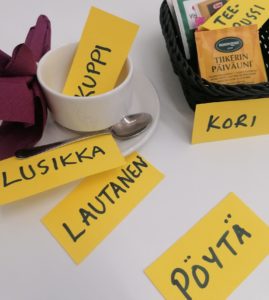 Languages can be learned in authentic environments through experiences. (ALT: Coffee mug, plate, coffee spoon, basket, teebags on a table and their Finnish translations written in small papers next to them).