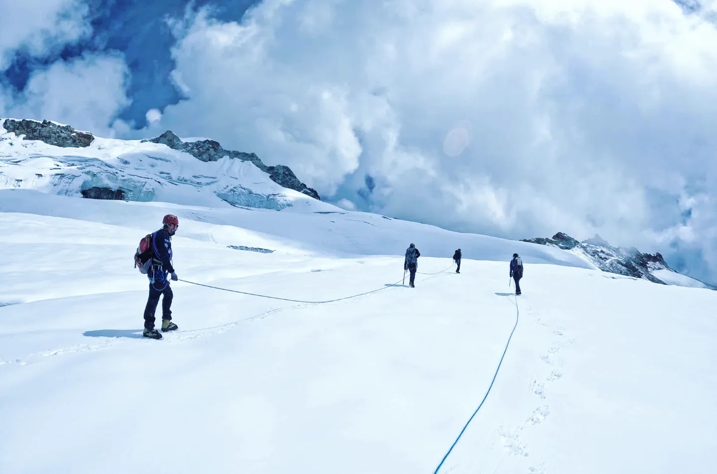Four people with backpacks and connected to each other with ropes, trekking up a snow-covered mountain with peaks in the backgroun