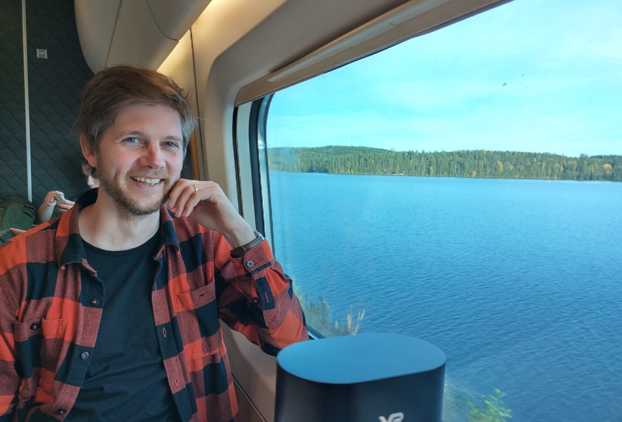 Kuvassa rennosti luontohenkisesti pukeutunut mies junassa, hymyilee kameralle ja nojaa kättään poskeen. Vieressä on ikkuna ja siitä näkyy sininen järvimaisema. Kuva on otettu junassa. 