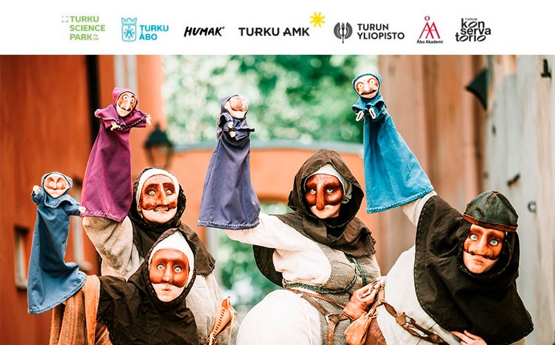 Kuvassa neljä naamiot kasvoillaan olevaa hahmoa vanhanaikaisissa puvuissa ulkoilmassa. Kuvan yläreunassa rivi Kulttuurikaupunki osallistujien logoja.