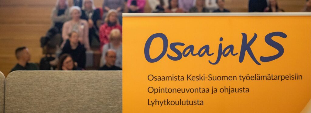 Yleisö on kokoontunut luentosaliin ja edessä rollup, jossa teksti OsaajaKS: Osaamista Keski-Suomen työelämätarpeisiin, opintoneuvontaa ja ohjausta sekä lyhytkoulutusta.
