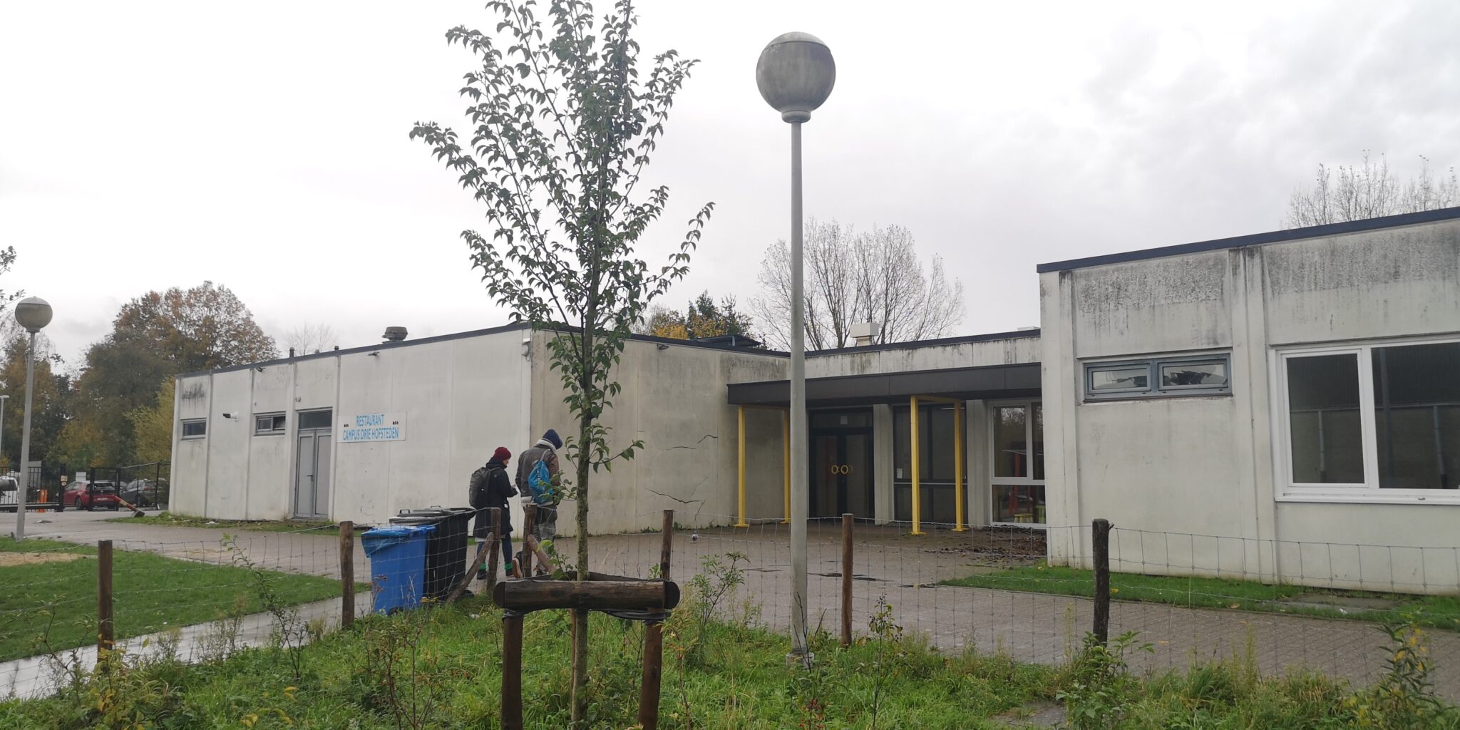 Kaksi henkilöä kävelee kohti harmaan koulurakennuksen sisäänkäyntiä sateisessa säässä.