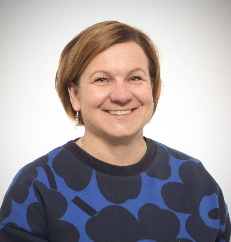 Alt-teksti: Hymyilevä keski-ikäinen nainen lyhyissä vaaleanruskeissa hiuksissa. Hänellä on yllään sininen pusero, jossa on suuria tummia kukkakuvioita, sekä pienet roikkuvat korvakorut. Taustalla on vaalea, neutraali studiotausta.