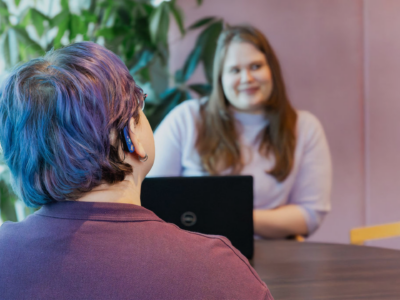 Two adults sit across a table in a bright office-like space; the foreground shows the back of a person with short blue-purple hair wearing a visible cochlear implant behind the ear, while a smiling woman with a laptop listens from the other side.