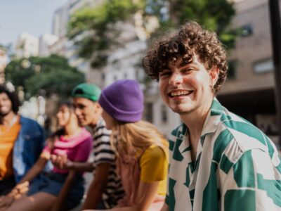 A group of young people sit outdoors in an urban setting on a sunny day. They are chatting casually on a bench, and the person in the foreground is wearing a green‑and‑white patterned shirt. Trees and buildings are visible in the background.
