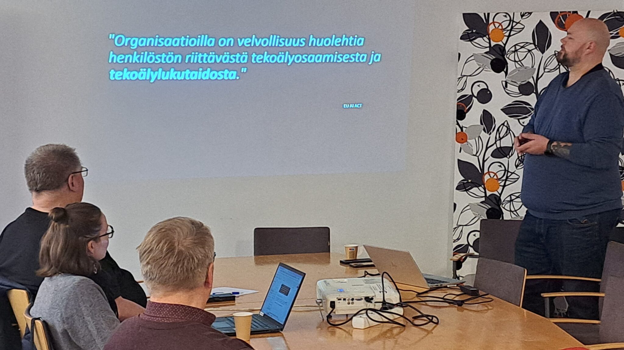 Kolme ihmistä on pöydän ääressä, selin kameraan. Pöydällä on kaksi kannettavaa tietokonetta ja projektori. Pöydän sivussa seisoo mies.