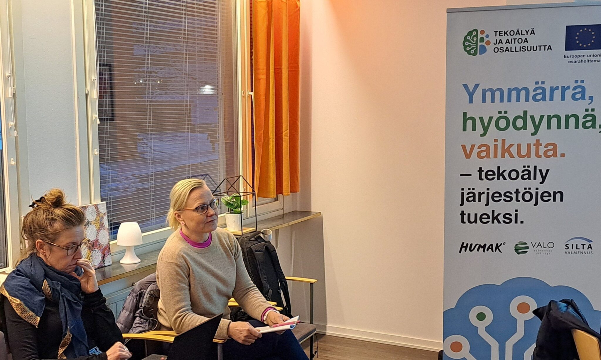  Kaksi naishenkilöä istuu tuoleilla, toisella kädessä lehtiö. Sivussa on hankkeen banderolli.