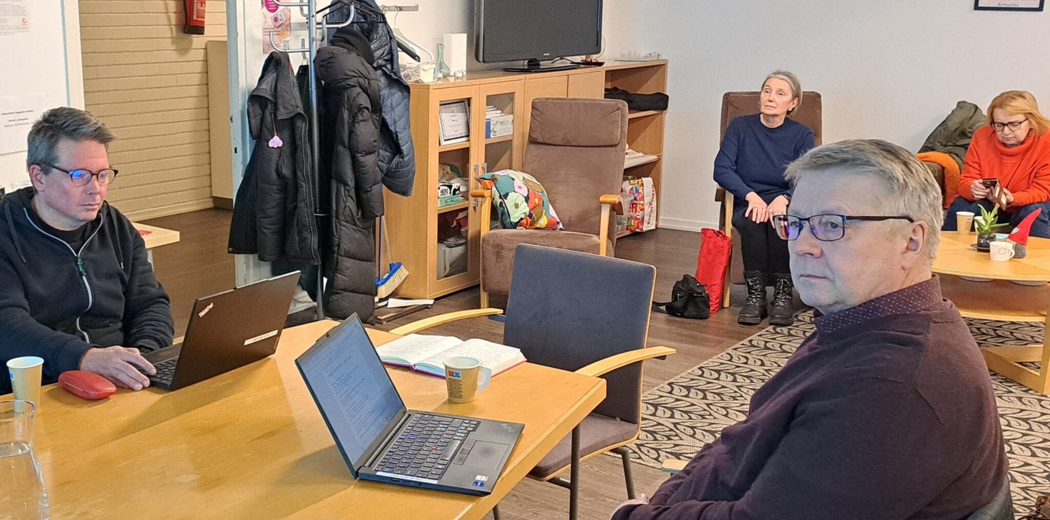 Kaksi miestä istuu pöydässä, pöydällä on kannettavat tietokoneet. Taustalla on kaksi naista nojatuoleissa, toisella matkapuhelin kädessään.