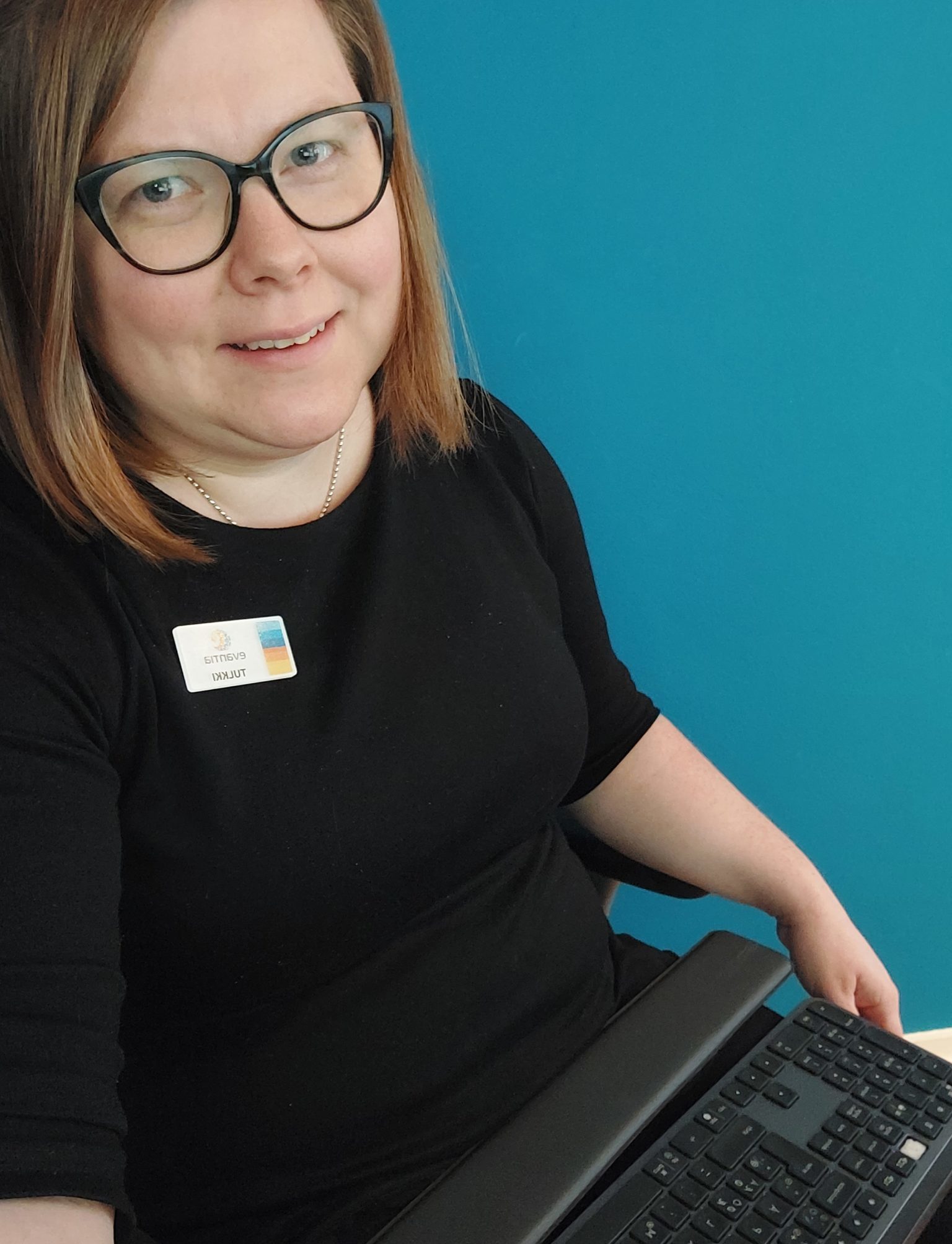 Hennalla on ruskeat hiukset, musta paita, silmälasit ja hymyilevät kasvot. Sylissään Hennalla on kirjoitustulkille tyypillinen näppäimistö.