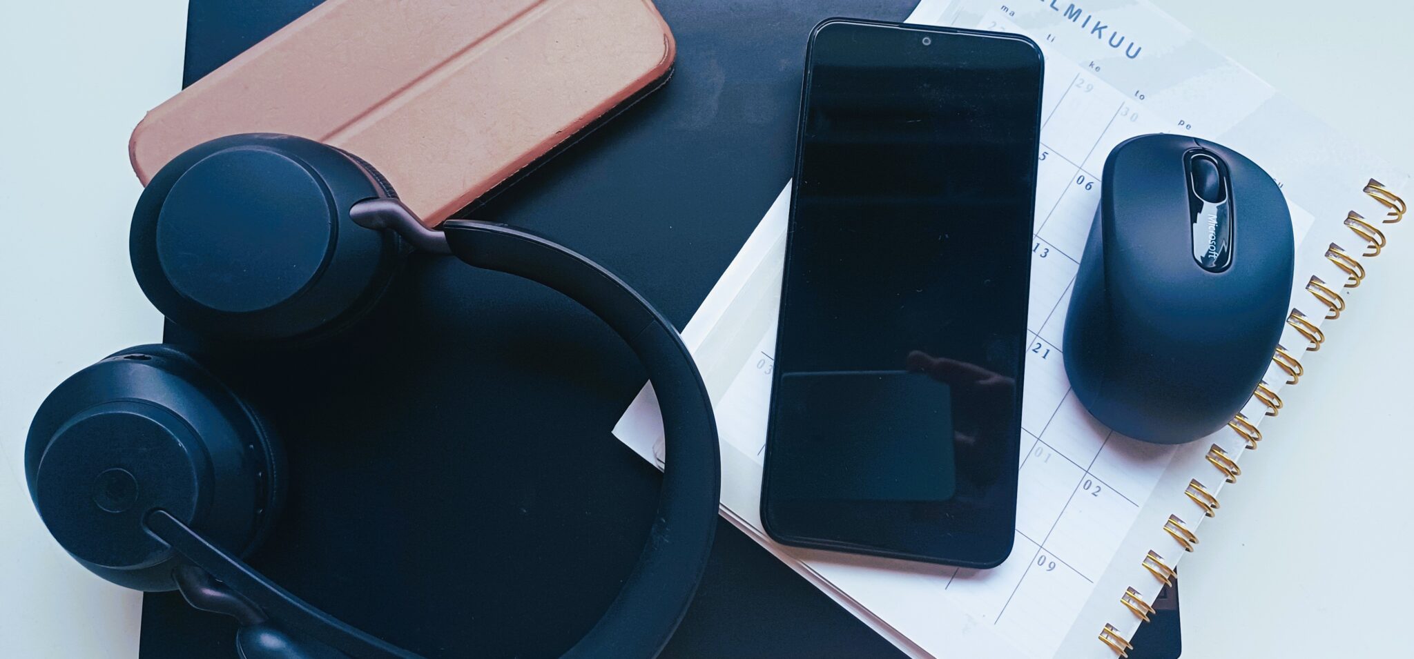 Työpöydällä on mustat kuulokkeet mikrofonilla, älypuhelin, langaton hiiri, ruskea tablettikotelo ja kalenteri.
