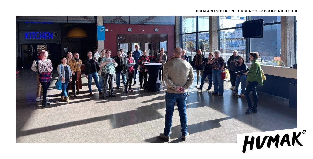 Mies seisoo selin kameraan päin suuressa aulassa, jonka suurista ikkunoista tulvii sisään valoa. Miehen edessä on noin 20 hengen ryhmä, joka näyttää kuuntelevan kiinnostuneena miehen puheita. 