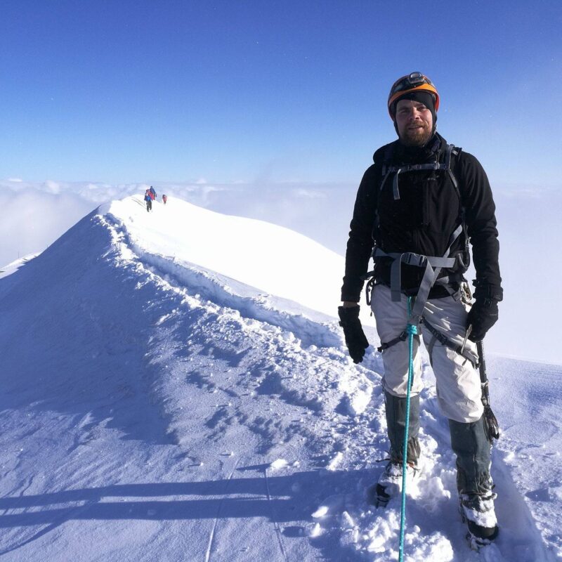 Hymyilevä mies lumivuoren huipulla vuorikiipeilijän varusteet päällä