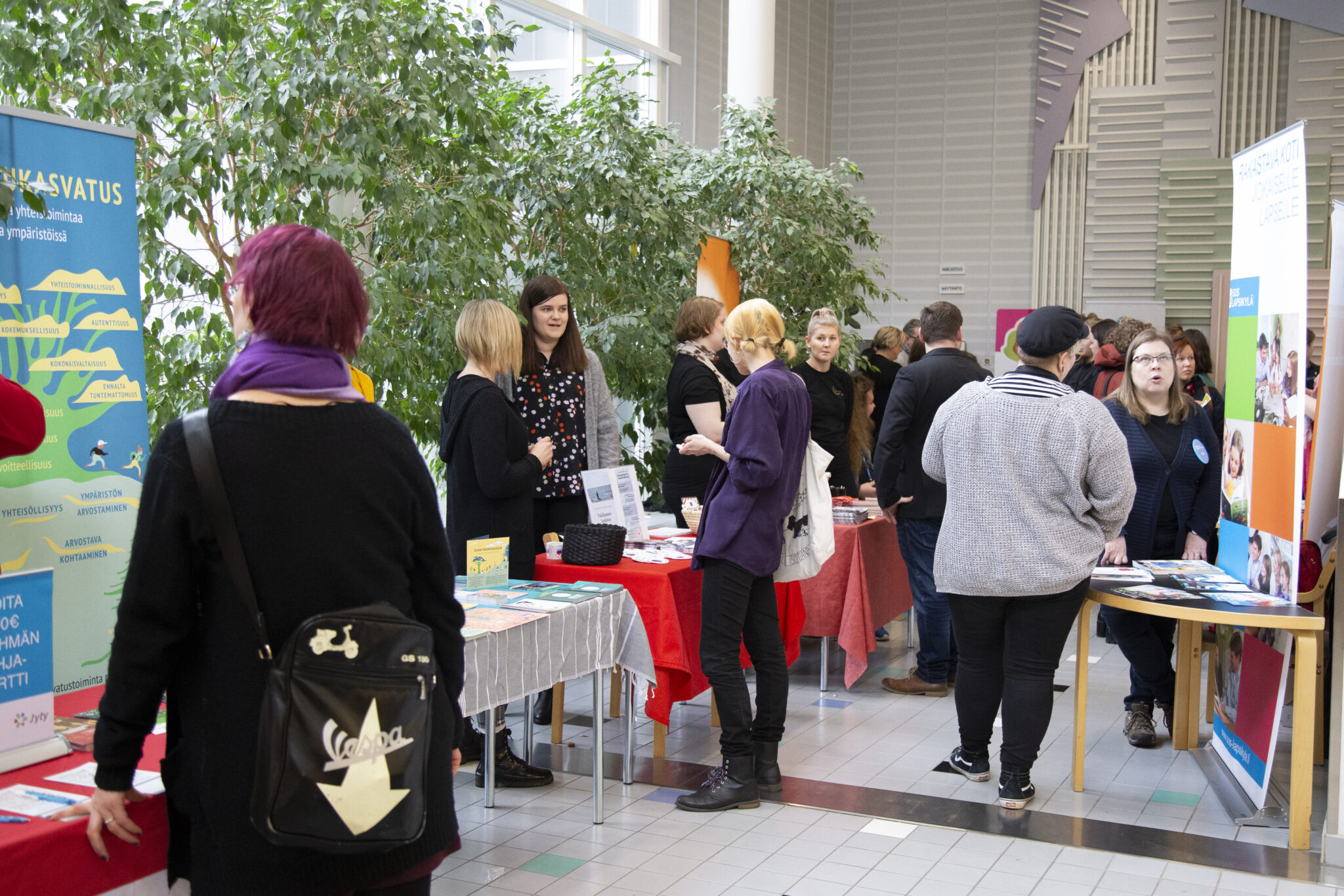 People are talking and exploring different information tables at an indoor event. The tables display brochures, posters, and other materials, with green plants and event banners visible in the background.