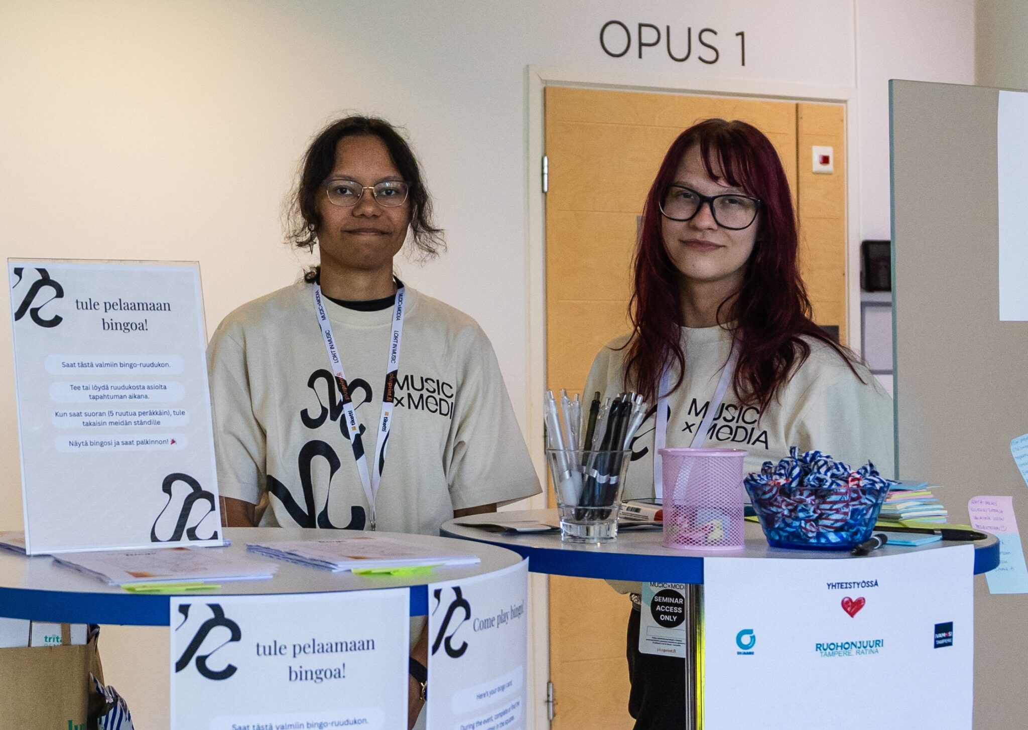 Kaksi nuorta seisoo vierekkäin pystypöytien takana ja katsoo kameraan.