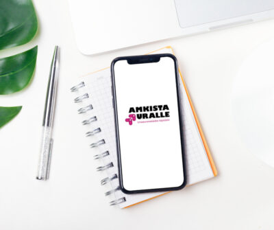Älypuhelin lepää kierresidonnaisen muistivihkon päällä, ja puhelimen näytöllä näkyy “AMKISTA URALLE” -logo. Vieressä on kynä, vihreä kasvinlehti ja osa kannettavaa tietokonetta.