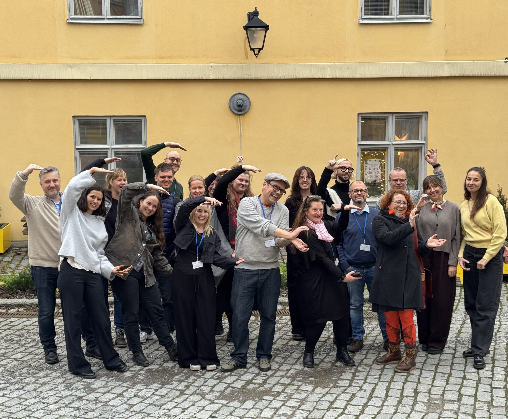 Ryhmä ihmisiä poseeraa ulkona keltaisen rakennuksen edessä. He seisovat rivissä ja muodostavat käsillään kaaria ja ympyröitä, hymyillen kameralle.