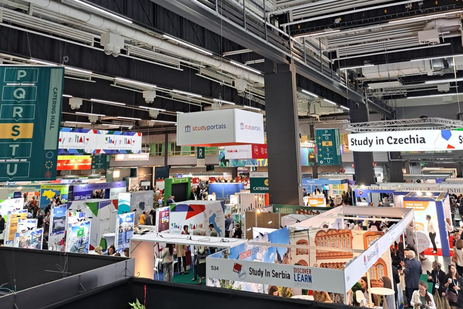 A busy international education fair inside a large exhibition hall, with booths from various countries including Italy, Serbia, and Czechia. Hanging signs display names like ‘Study in Italy’, ‘Study in Czechia’, and ‘Studyportals’, while attendees walk between colorful stalls.