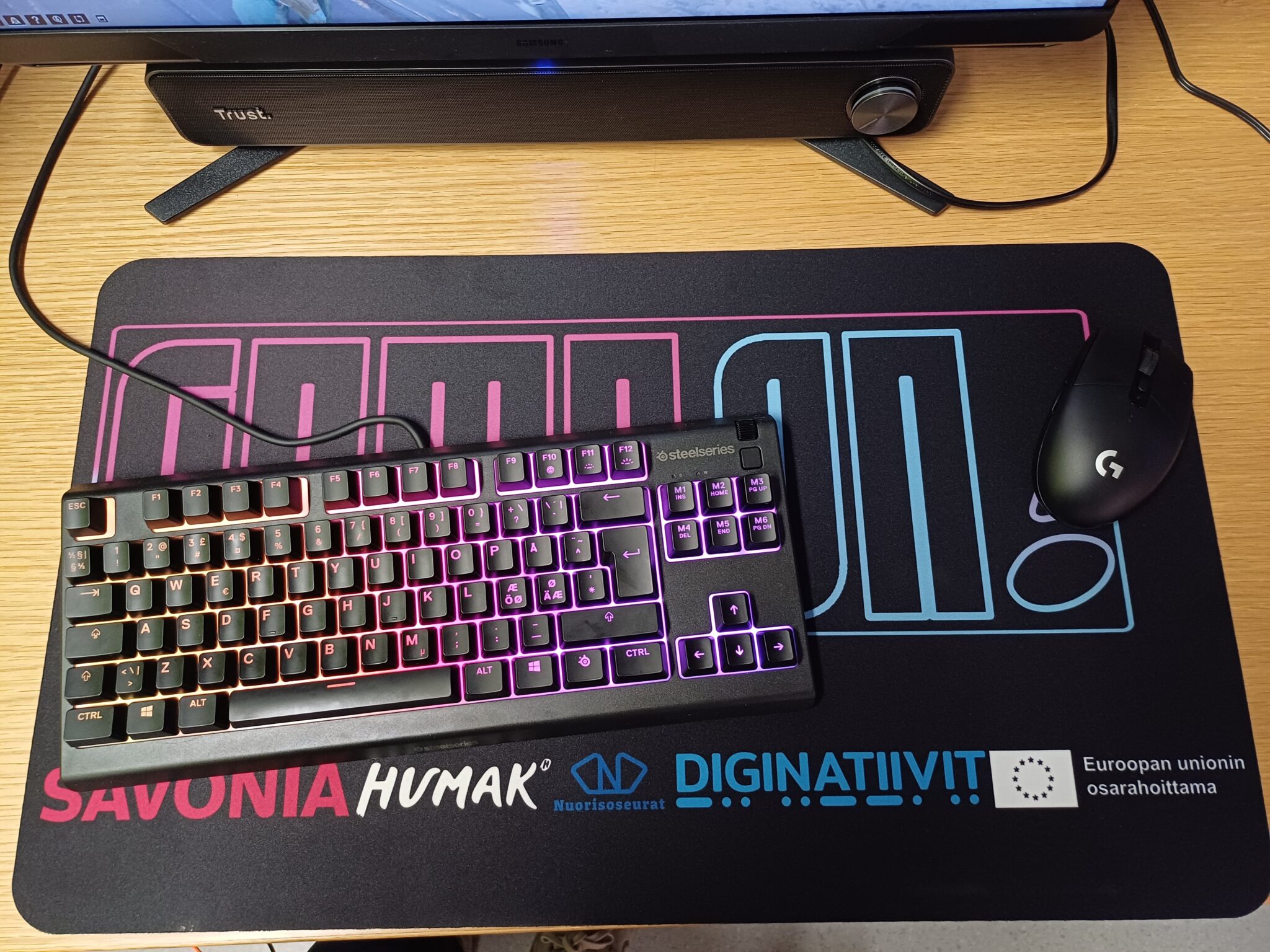 Lähikuva pelihiirimatosta, jonka päällä on värivaloin valaistu näppäimistö ja hiiri. Hiirimatossa lukee "GAMEON" ja siinä ovat logot: Savonia, Humak, Nuorisoseurat, Diginatiivit sekä Euroopan unionin tunnus ja teksti "Euroopan unionin osarahoittama".