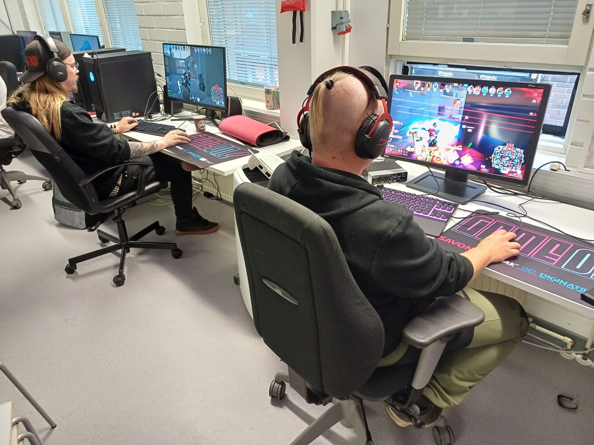 Kaksi henkilöä istuu tietokoneilla pelihuoneessa pelaten tietokonepeliä kuulokkeet päässä. Molemmilla on suuri hiirimatto, jossa lukee "GAMEON" sekä logoja, kuten Savonia, Humak, Diginatiivit ja Euroopan unionin tunnus.