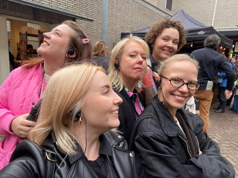 Kuvassa viisi henkilöä, joilla kaikilla on toisella korvallaan musta johdollinen kuuloke. Muut henkilöt hymyilevät leveästi, mutta keskellä olevalla henkilöllä on viekas ilme.