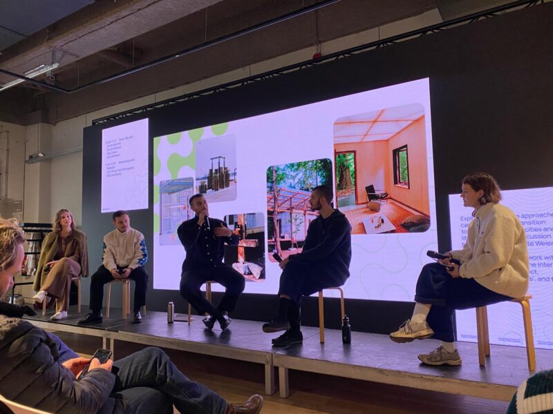 Kuvassa matala lavarakenne, jonka päällä istuu viisi paneelissa keskustelevaa henkilöä jakkaroilla. Heidän takanaan on valkokangas/näyttö, jossa on kuvia Gigi Totaron töistä. Totarolla on kädessään mikrofoni, johon hän puhuu