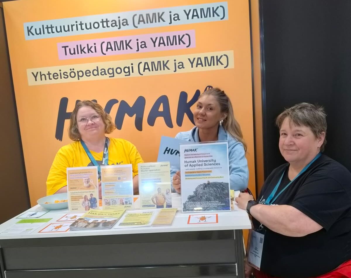 Humakin ständi rekrymessuilla, taustaseinä ja pöytä, joiden välissä kolme naista. 