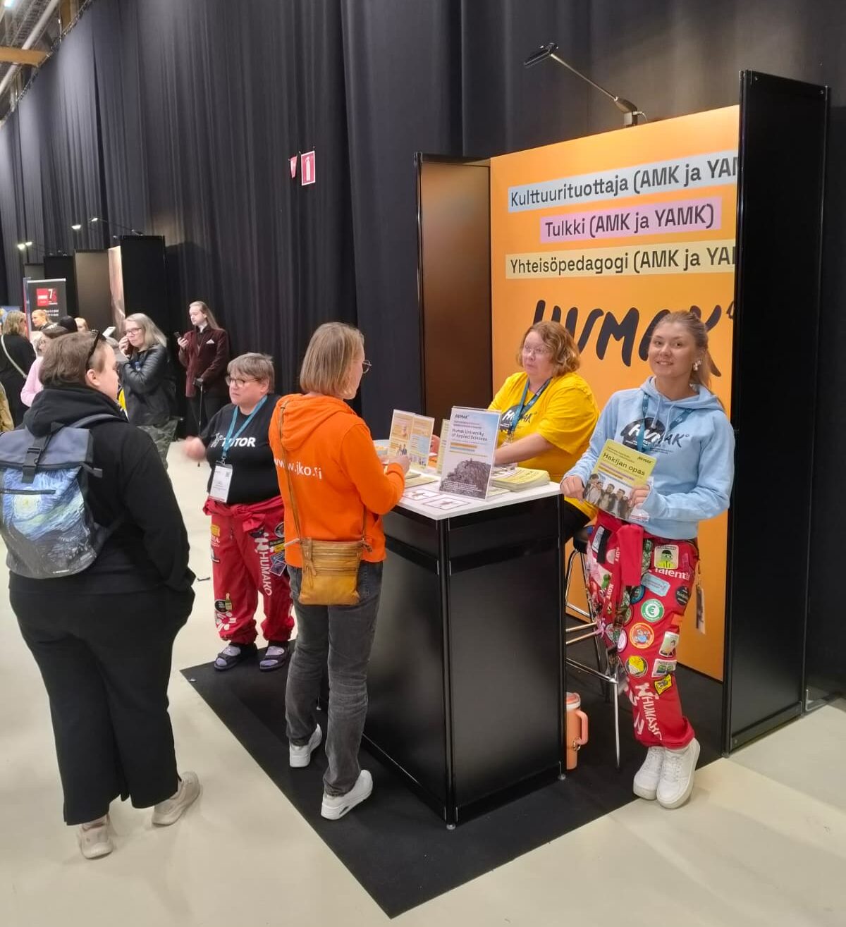 Kuvassa Humak-esittelypiste messuilla. Kolme naista seisoo esittelypisteen pystymallisen pöydän ympärillä, pöydällä esitteitä ja takana Humak-brändätty taustasermi. 
