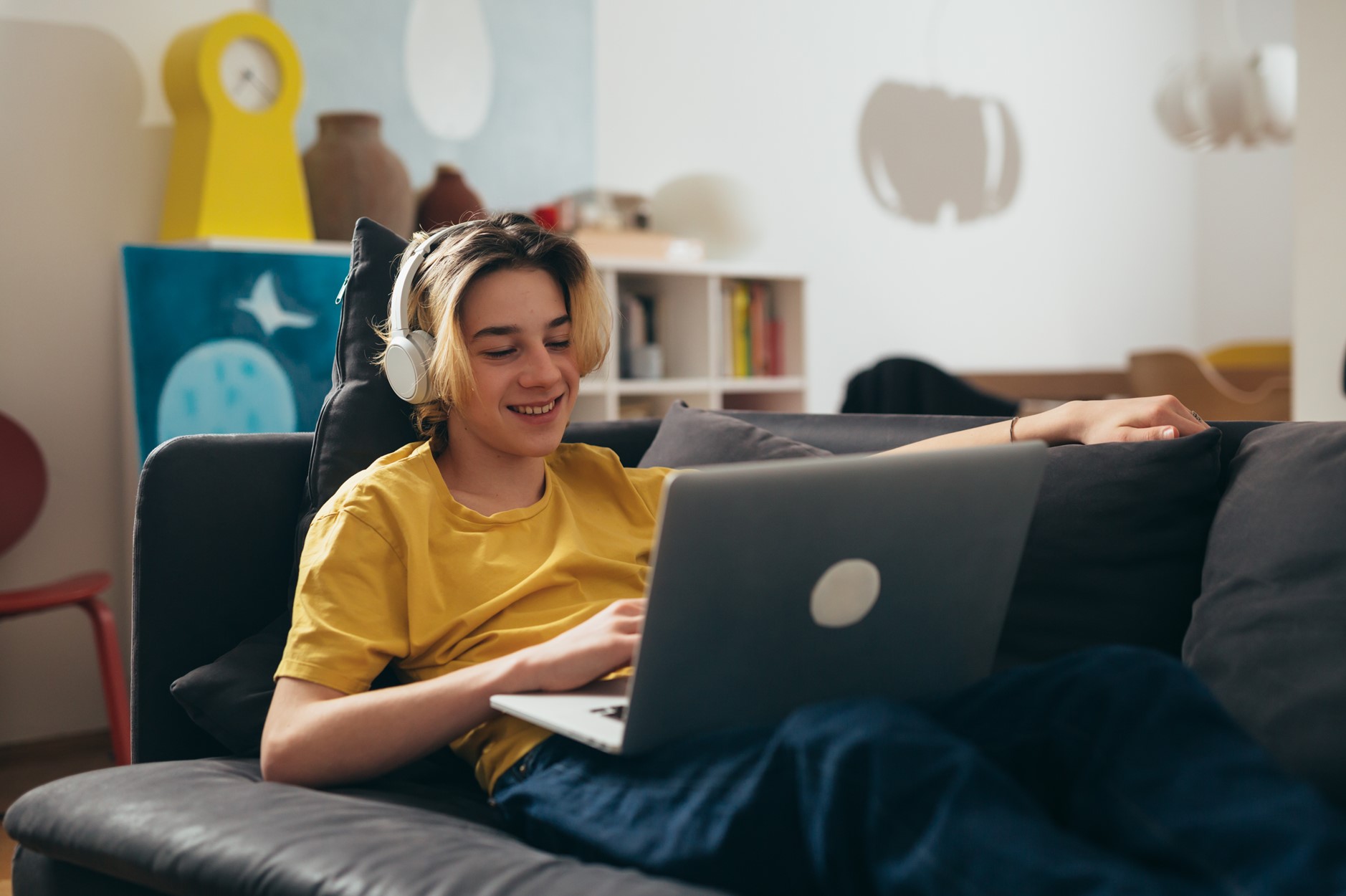 Nuori istuu sohvalla kannettava tietokone sylissään, kuulokkeet päässä ja keltainen paita yllään. Taustalla näkyy keltainen kello, keramiikkaesineitä sekä kirjahyllyjä kirjoineen ja muine tavaroineen.