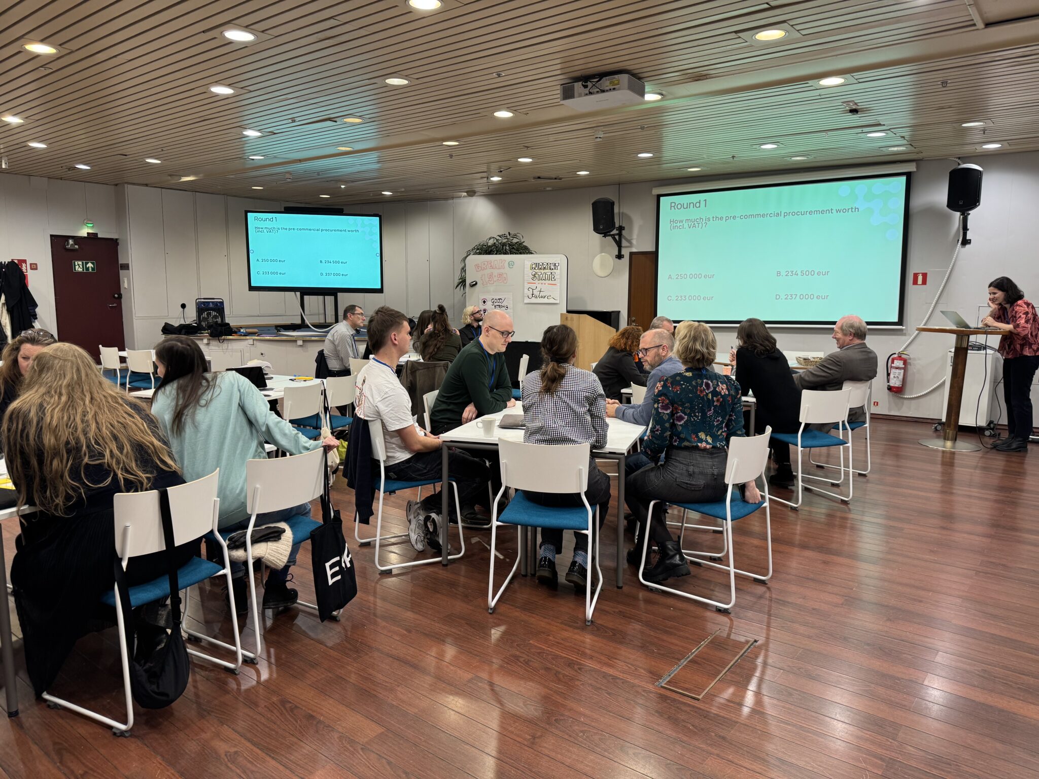 Joukko ihmisiä istuu pöytien äärellä. Kaksi näyttöä heijastavat dia, jossa lukee " Rpund 1".