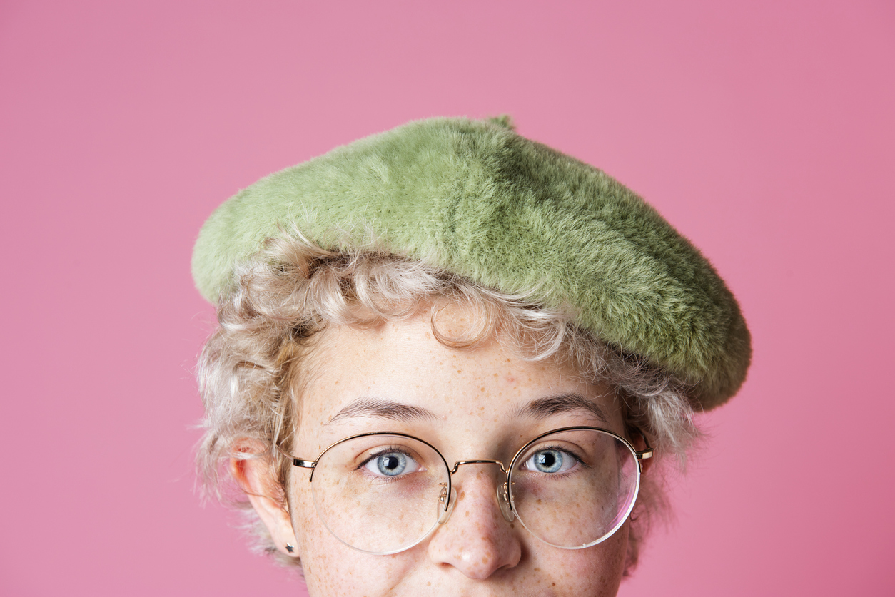 Kuvassa näkyy baskeripäisen naisen kasvojen yläosa. Naisella on silmälasit. Tausta on vaaleanpunainen.