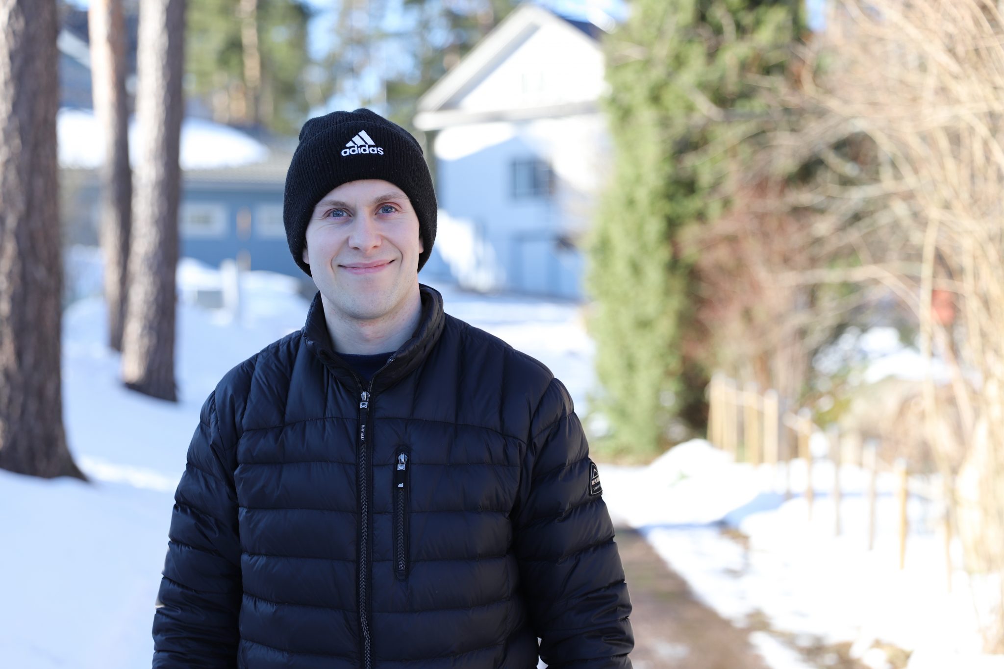 Mustaan pipoon ja takkiin pukeutunut mies hymyilee talvisessa maisemassa.